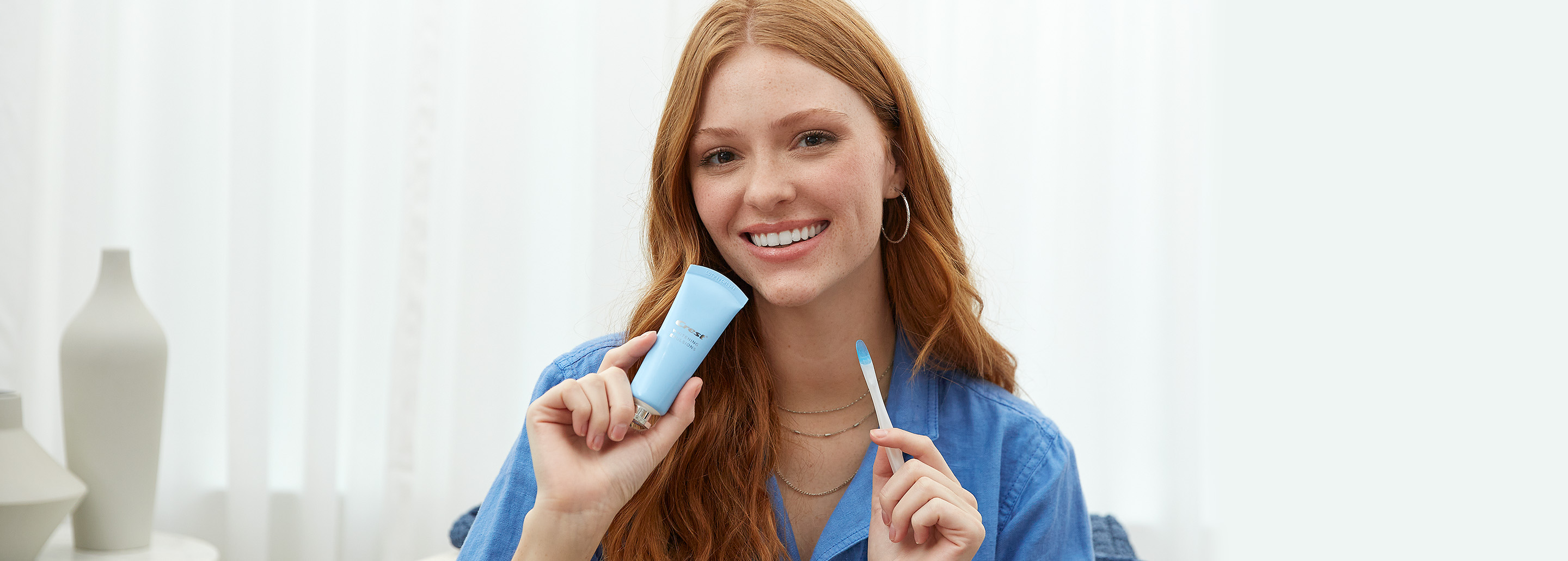 Redhead woman smiles and holds Crest Whitening Emulsions applicator wand and treatment tube. 
