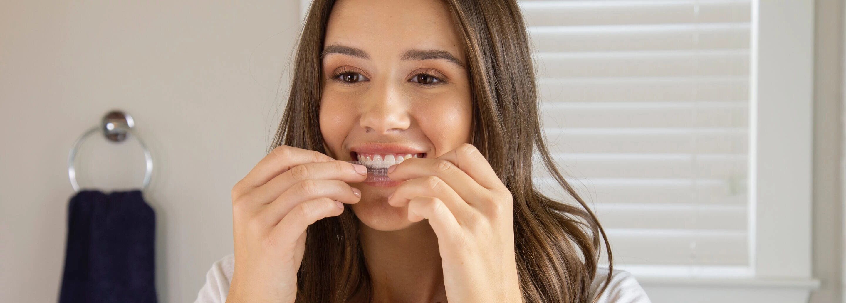 Young brunette woman smiles as she applies Crest 3DWhitestrips to her teeth.